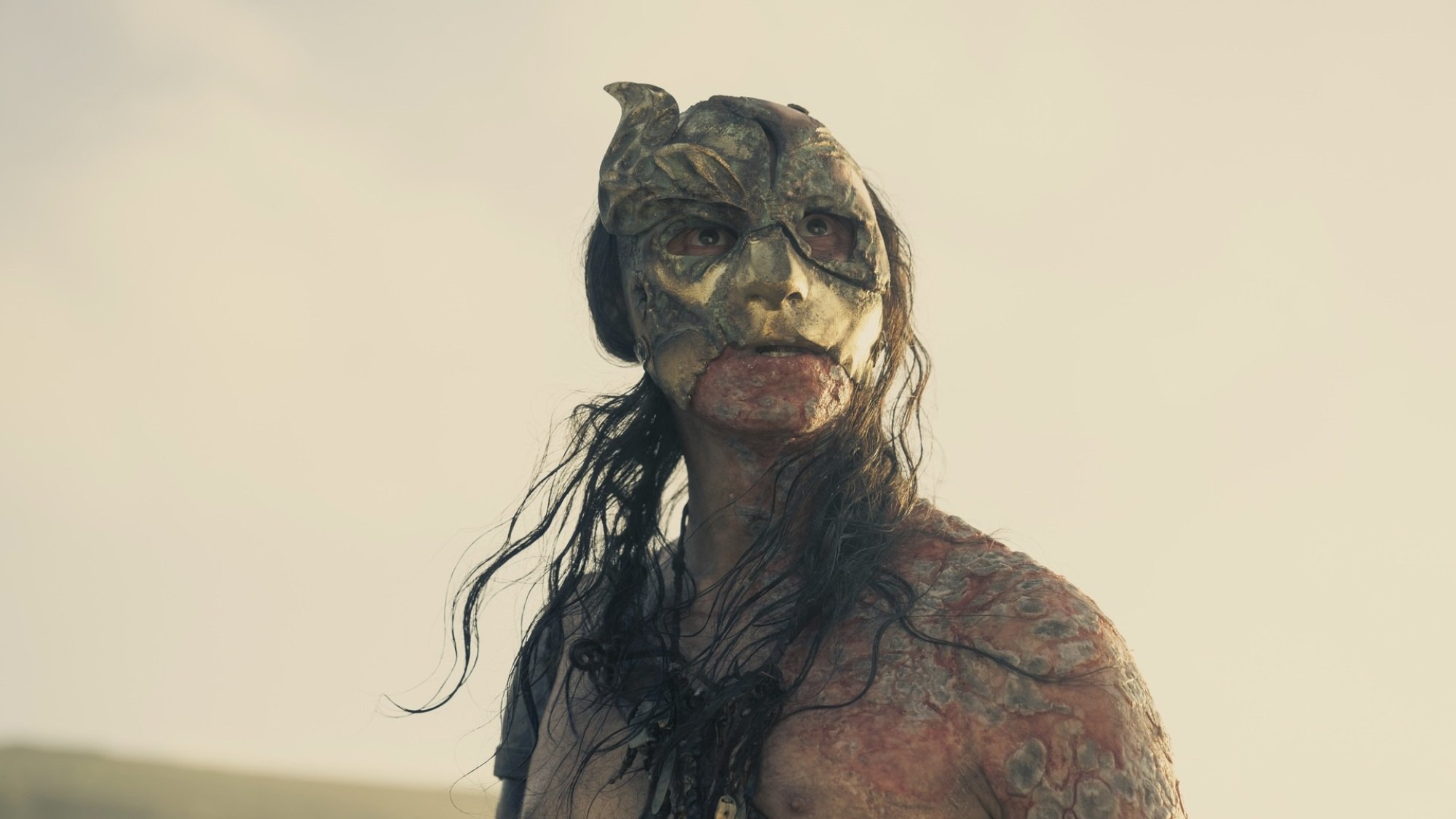 A badly sunburned man with long brown hair and a cracked golden mask.