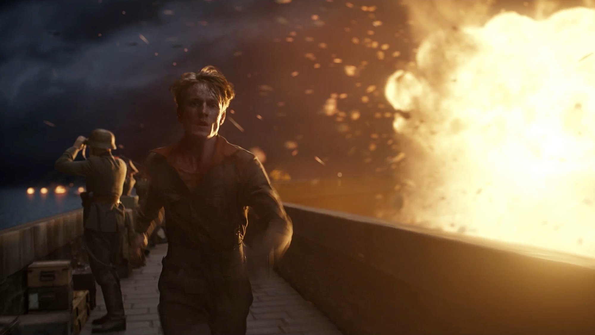 A young man in a soldier's uniform runs along a wall as an explosion occurs nearby.