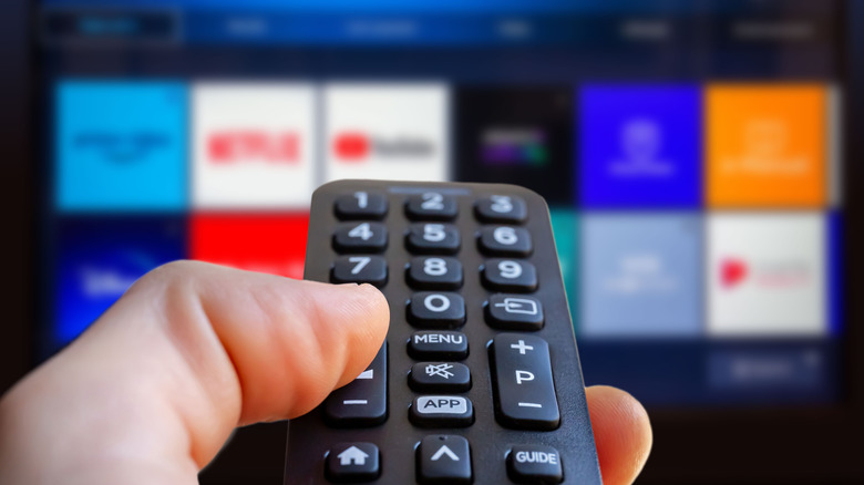 Hand holding a remote with a blurred out TV showing streaming apps in the background.
