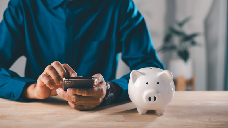 A person sitting next to a piggy bank while using their mobile device.